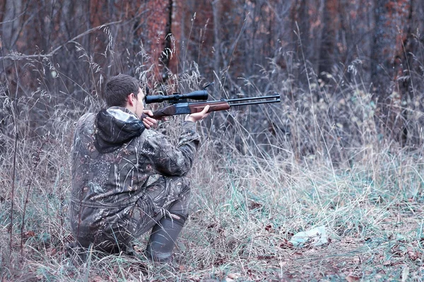 En man i kamouflage och med ett jaktgevär i en skog på en sp — Stockfoto