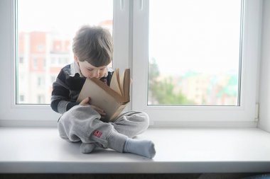Küçük çocuk bir kitap okuyor. Çocuğun pencerenin önünde oturur bir