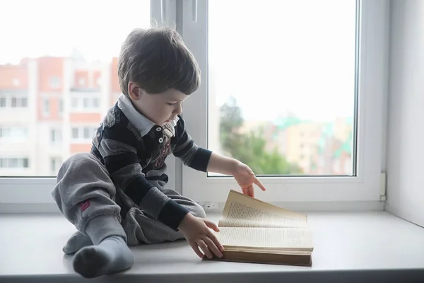 Het jongetje is het lezen van een boek. Het kind zit bij het raam een — Stockfoto