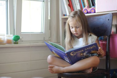 Küçük kız odada oturuyor ve kitap okuyor.