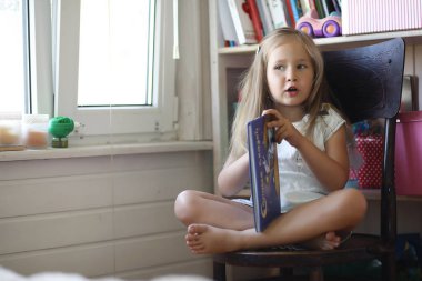 Küçük kız odada oturuyor ve kitap okuyor.