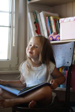 Küçük kız odada oturuyor ve kitap okuyor.