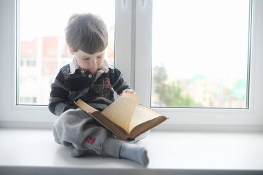 Küçük çocuk bir kitap okuyor. Çocuğun pencerenin önünde oturur bir