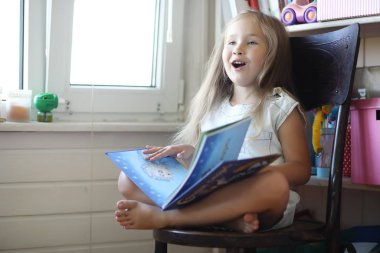 Küçük kız odada oturuyor ve kitap okuyor.