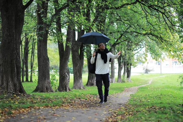 En ung man i glasögon går i parken med ett paraply under — Stockfoto