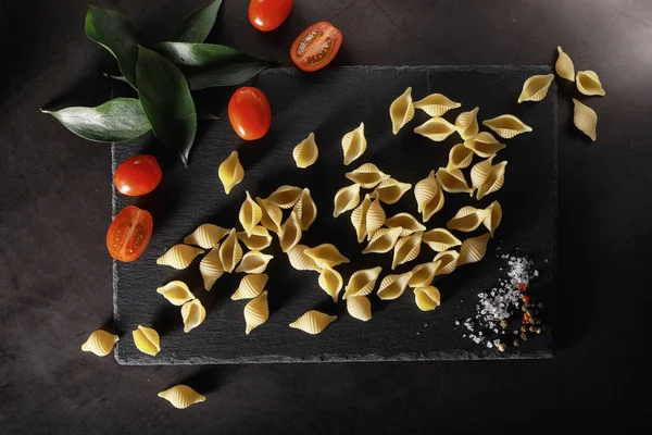Cáscaras de pasta sobre piedra negra. Tomates y hojas de laurel con allspi — Foto de Stock