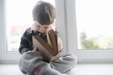 Küçük çocuk bir kitap okuyor. Çocuğun pencerenin önünde oturur bir