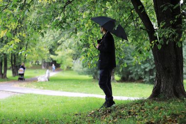 Gözlüklü genç bir adam parkta şemsiyeyle yürür.