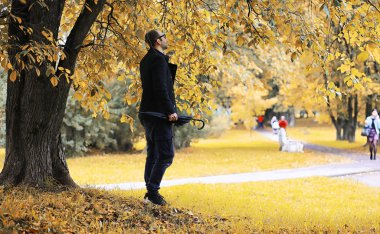 Gözlüklü genç bir adam parkta şemsiyeyle yürür.