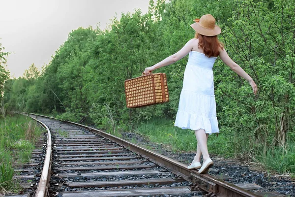 Beyaz elbiseli ve hasır bavullu bir kız raylarda yürüyor. — Stok fotoğraf