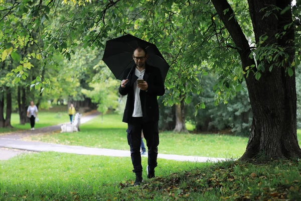 Egy szemüveges fiatalember besétál a parkba egy esernyővel. — Stock Fotó