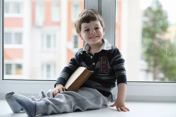 Het jongetje is het lezen van een boek. Het kind zit bij het raam een — Stockfoto