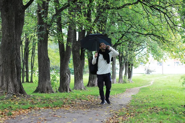 Egy szemüveges fiatalember besétál a parkba egy esernyővel. — Stock Fotó