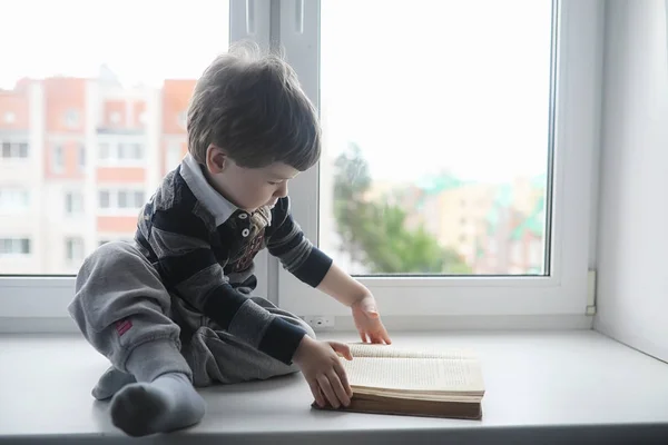 Küçük çocuk bir kitap okuyor. Çocuğun pencerenin önünde oturur bir — Stok fotoğraf