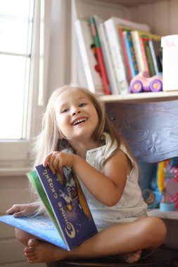 Küçük kız odada oturuyor ve kitap okuyor.