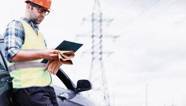 Kasklı ve üniformalı bir adam, sahada bir elektrikçi. Profesyonel elektrik mühendisi elektrik hatlarını inceliyor..