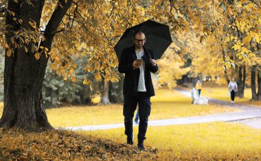 Gözlüklü genç bir adam yağmur sırasında elinde şemsiyeyle parkta yürür..