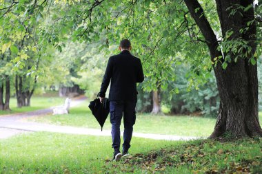 Gözlüklü genç bir adam yağmur sırasında elinde şemsiyeyle parkta yürür..