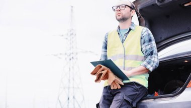 Kasklı ve üniformalı bir adam, sahada bir elektrikçi. Profesyonel elektrik mühendisi elektrik hatlarını inceliyor..