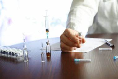 Bir adam tıbbi belge imzalıyor. Tıbbi ekipmanlar masada. Stetoskop ve şırıngalı ampuller. Ofiste notlar alıyor. Tıp Merkezi.