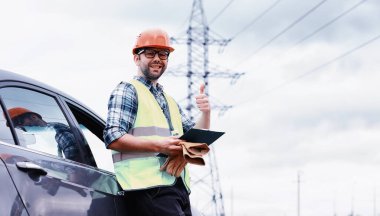 Kasklı ve üniformalı bir adam, sahada bir elektrikçi. Profesyonel elektrik mühendisi elektrik hatlarını inceliyor..