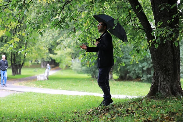 一个戴眼镜的年轻人在下雨时带着雨伞在公园里散步 — 图库照片