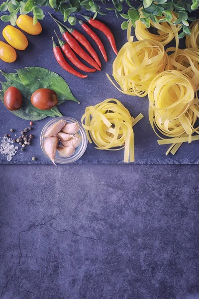 Pasta on the table with spices and vegetables. Noodles with vegetables for cooking on black stone background.