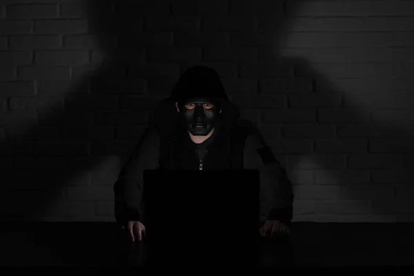 Hacker in black mask and hood at the table in front of the monito
