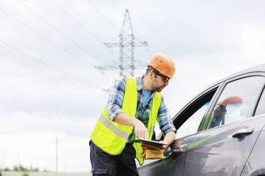 Kasklı ve üniformalı bir adam, sahada bir elektrikçi. Profesyonel elektrik mühendisi elektrik hatlarını inceliyor..