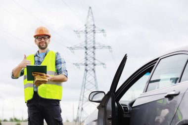 Kasklı ve üniformalı bir adam, sahada bir elektrikçi. Profesyonel elektrik mühendisi elektrik hatlarını inceliyor..