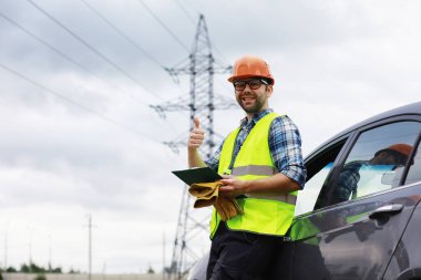 Kasklı ve üniformalı bir adam, sahada bir elektrikçi. Profesyonel elektrik mühendisi elektrik hatlarını inceliyor..