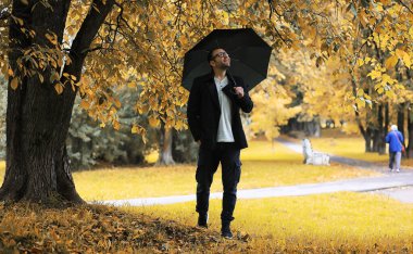 Gözlüklü genç bir adam yağmur sırasında elinde şemsiyeyle parkta yürür..