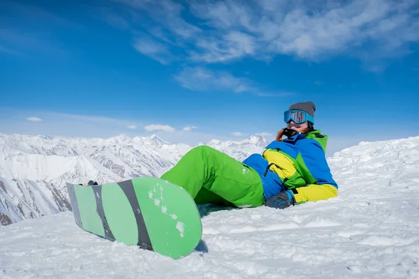 Uma menina em um terno de esqui jaz na neve com um snowboard vestido um — Fotografia de Stock