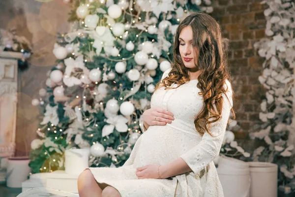 Pregnant woman holding a belly near a Christmas tree with lights — Stock Photo, Image