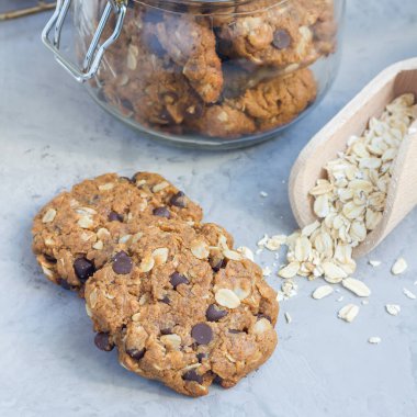 Unsuz glütensiz fıstık ezmesi, yulaf ezmesi ve çikolata parçalı kurabiyeler cam kavanozda ve masada, kare formatta.
