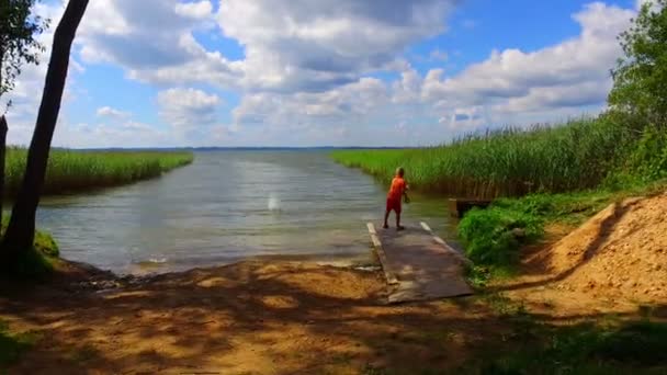 Мальчик играет на зеленом берегу озера — стоковое видео