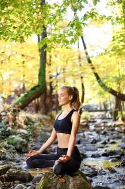 Yoga yapan güzel bir kız, spor yapan, parkta meditasyon yapan, doğa geçmişi olan bir orman. Spor konsepti. Parkta sağlıklı yaşam tarzı eğitimi. Nehir kenarında spor yapan bir kız. Lotus yogası.