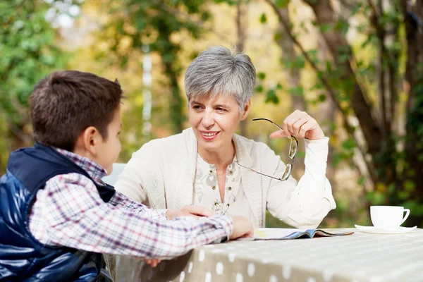 Older woman with grandson in the park reading a book, drinking coffee and talking — Stock Photo, Image