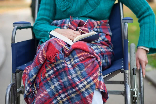 Una donna anziana in sedia a rotelle che legge un libro in un parco con colori autunnali — Foto Stock