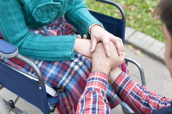 мужские руки, держащие за руки женщину, которая сидит в инвалидном кресле
