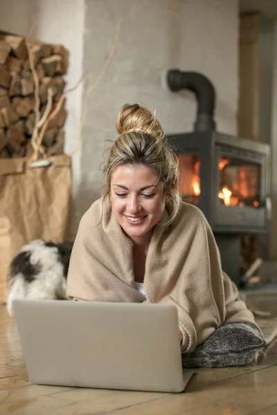 Young beautiful woman in the living room sits on the floor and communicates with friends over the computer, covered with a blanket for the cold in the back of the fireplace