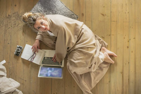 Young beautiful woman in the living room lying on the floor and communicates with friends over the computer, covered with a blanket for the cold. Top view.