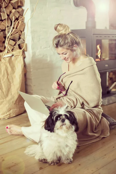 Young beautiful woman in the living room sits on the floor and communicates with friends over the computer, covered with a blanket for the cold in the back of the fireplace