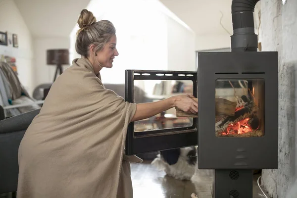 Beautiful middle-aged woman inserts wooden logs into the fireplace. Living room with fire.