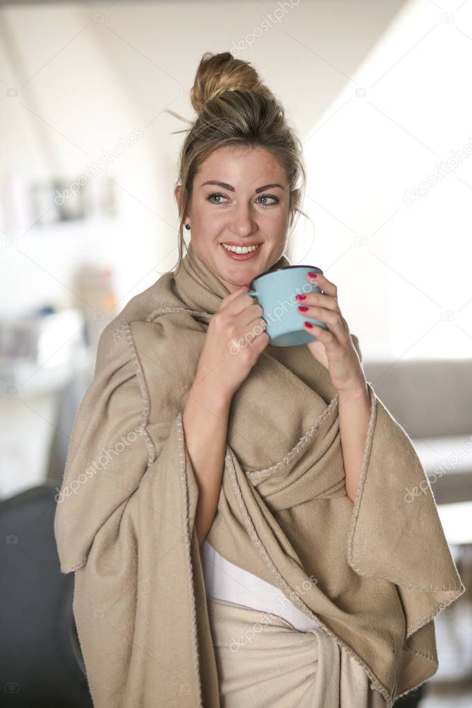 Beautiful middle-aged woman drinks tea from a glass in a cold living room dressed in a cotton becouse of cold winter. A sick girl drinks from a blue cup in the living room.