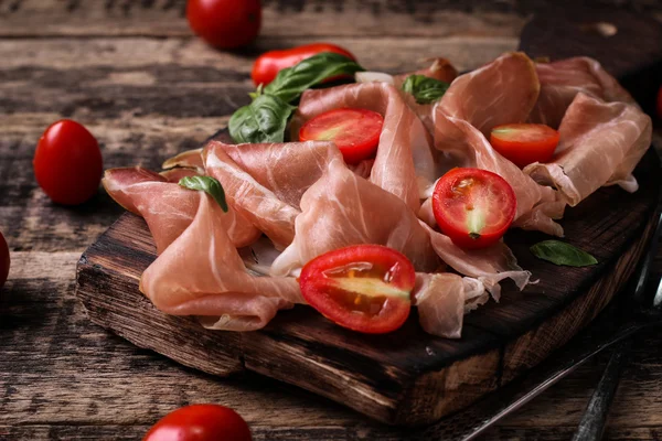 Thin slices of prosciutto with mixed with basil, cherry tomatoes on wooden cutting board,vintage background — Stock Photo, Image