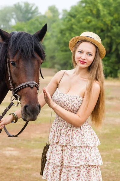 Beautiful girl in a straw hat and summer dress posing with a horse — Stock Photo, Image
