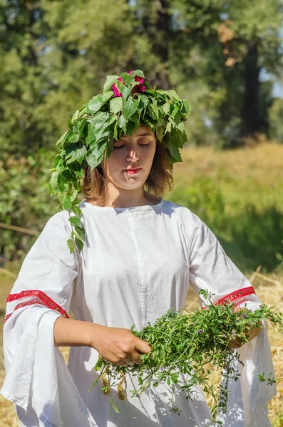 Girl in Russian folk clothes, weaves a wreath of grass and leaves — Stock Photo, Image