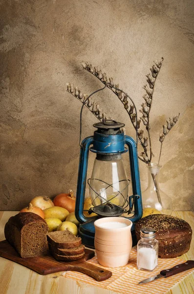 Milk in ceramic ware, rye bread, salt shaker with salt, vegetables, dry henbane branch in a decanter and kerosene lantern on a wooden table. Still-life in the rustic style