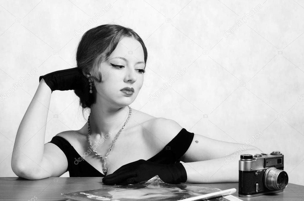 Beautiful girl in evening dress and long gloves, sitting at the table with magazines and a camera. Black and white portrait in retro style